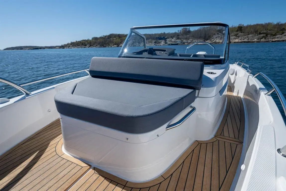A grey cushioned seating area on a boat, with wooden decking and calm water in the background under a clear blue sky.